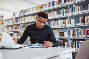 In stylish black shirt. African american man sitting in the library and searching for some information in the books photo