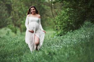 Going forward and smiling. Beautiful pregnant woman in dress have a walk outdoors. Positive brunette photo