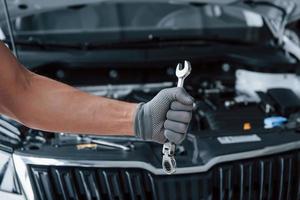 Blurred background. Man's hand in glove holds wrench in front of broken automobile photo