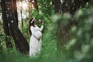 luz del sol y flores. hermosa mujer embarazada vestida tiene un paseo al aire libre. morena positiva foto