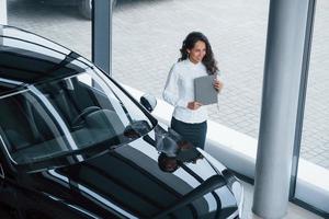 Near the windows. Curly haired female manager stands near the car in automobile salon photo