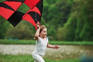 It feels awesome. Positive female child running with red and black colored kite in hands outdoors photo