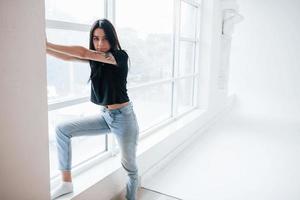 de pie en el alféizar de la ventana. Una joven adolescente morena tiene una sesión de fotos en el estudio durante el día