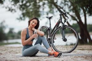 Water is very important. Female cyclist with good body shape sitting near her bike on beach at daytime photo