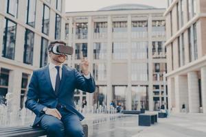 Smiling businessman with stubble sitting on bench in town square with VR glasses on photo