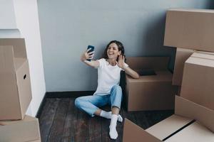 Happy young spanish woman in jeans and white t-shirt unpacking boxes having video call on phone. photo