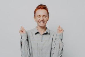 Excited overjoyed young ginger female raising hands with clenched fists, keeping eyes closed photo