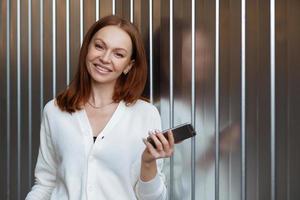 Satisfied female entrepeneur with toothy smile, wears white jumper, holds modern cell phone, sends text message, waits for business partner, stands indoor with free space for your promotion. photo