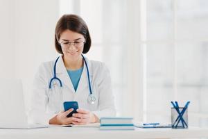 Female medical worker uses smartphone, reads advice how to cure disease in internet, poses in medical office, wears special medical uniform, sees results of examination for patient, examines chart photo