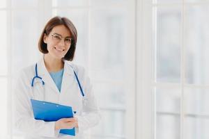 Good looking female expert creats medical prescription, writes down information in clipboard, thinks about good advice for patient, stands indoor, copy space for your information. Medicine concept photo