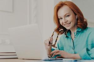 Smiling good looking female employee talks on video call, has web conference with colleagues, watches webinar on modern laptop, poses in coworking space. Student learns foreign languages online photo