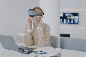 Excited girl in VR headset sitting at the desk in front of laptop. Working on project in cyberspace. photo