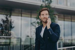 Male entrepreneur talks via phone solves urgent problems poses near business center photo