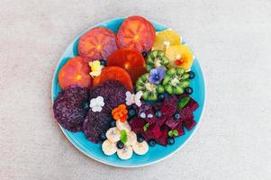 Top view of tropical fruit assortment on plate against white background. Fresh slices of dragon fruit, pitaya, kiwi, banana, blueberry decorated with small pansy. Bright colored healthy fruit photo