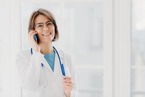 Positive woman doctor consults patient via cellphone, looks gladfully at camera, holds pen, wears white coat, poses in medical office, poses near window. Healthcare and consultation concept. photo