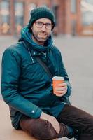 Stylish Caucasian man has free time, poses outside with disposable cup of coffee, dressed in hat and jacket, enjoys good rest, waits for someone at street, has satisfied expression. Lifestyle concept photo