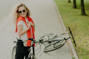 Cheerful curly haired female stops during way, poses on bicycle at road, friends bike lies near, wears sunglasses, t shirt and trousers, carries little bag around waist, being fit and healthy photo