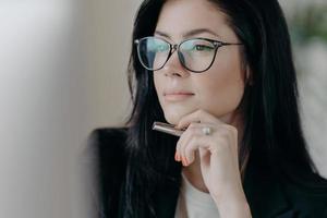 Serious woman author works on new publication, concentrated in display of computer, keeps pen in hand, wears spectacles for eye correction. Ambitious successful female entrepreneur manages business photo