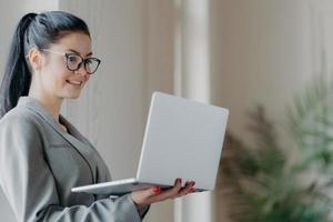 toma interior de un trabajador corporativo ocupado posa en un espacio de coworking, sostiene una computadora portátil moderna, transmite la fecha, está conectado a Internet inalámbrico, usa anteojos para la corrección visual, envía comentarios foto