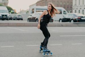 Sideways shot of active young woman in good physical shape rollerblades along city streets looks happily behind wears black sportsclothes leads healthy lifestyle. Recreation and hobby concept photo