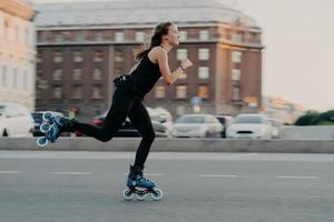 mujer atlética monta en rodillos se mueve muy rápido vestida con ropa activa disfruta de patinar siendo fotografiada en poses de acción en un lugar urbano dedicado a deportes extremos. concepto de estilo de vida activo foto