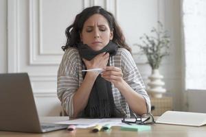 Worried young hispanic woman office worker with fever sitting workplace and looking at thermometer photo