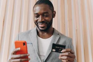 feliz hombre de negocios africano en traje con tarjeta de crédito y teléfono móvil mientras hace compras en línea foto