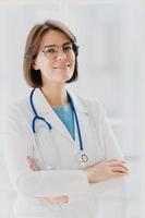 Shot of confident brunette female doctor or surgeon stands with hands crossed, wears white uniform, cares about health of patients ready to give advice any time. People, occupation, profession concept photo