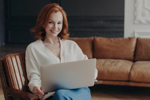 Gorgeous young ginger woman in white jumper sits on armchair with laptop computer, prepares for online interview with new employee, big comfortable sofa in background. Working distantly from home photo