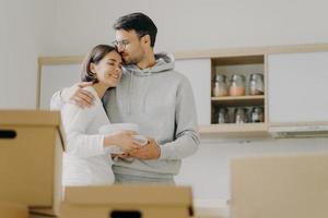 Young couple in love embrace and kiss with tender, hold pile of white plates, stand in kitchen during moving day, surrounded with many carton boxes filled with personal belongings, unpack stuff photo