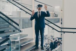 Businessman in VR headset stands on staircase in office lobby with hands up in air photo