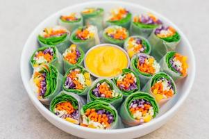 Vegan food concept. Tasty green spring rolls made of rice paper and spinach, filled with fresh chopped vegetables. Small plate with yellow curry sauce in middle photo