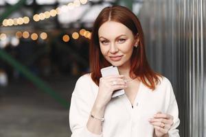 Photo of prosperous female business worker has break after work, sends text messages, holds cell phone in hand, dressed in white shirt, has browh hair, stands outdoor against blurred background