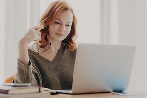 Concentrated female student reads information and checks data, focused at laptop screen, uses wireless internet connection, prepares for exam, poses at workplace, holds eyewear for vision correction photo