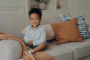 Sweet afro kid boy in casual clothes sitting on comfortable sofa and playing indoors photo