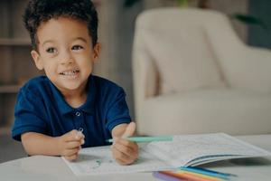 Cute afro american small boy in dark blue polo tshirt painting coloring book at home photo