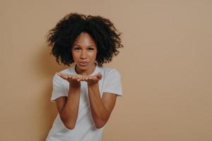 Beautiful curly flirty african woman sending air kiss and smiling while posing on beige background photo