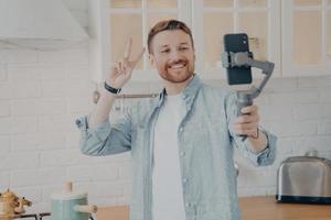 apuesto joven brunete sonriente con rastrojo haciendo selfie en la cocina foto