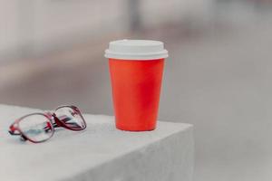 Horizontal shot of red coffee to go and optical glasses. Aromatic coffee for you to drink. Drinking concept. Paper cup of hot beverage photo