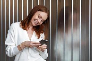 Positive successful European female economist makes money payment on website, poses with cell phone, dressed in white outfit, smiles happily, sends text message, enjoys online communication. photo