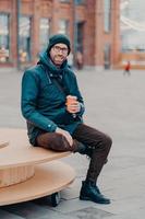 Concept of drinks and lifestyle. Carefree satisfied European man with bristle enjoys sunny day and hot beverage, smiles happily, sits against street background. Outdoor shot of handsome hipster photo