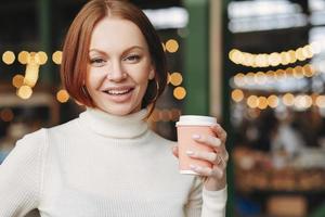 Optimistic lovely woman with dyed hair, satisfied expression, wears turtleneck jumper, holds takeaway coffee, poses in outdor restaurant, has nice beverage, being in high spirit, likes cappuccino photo