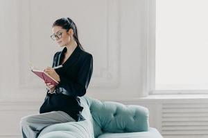 Attracative successful businesswoman in elegant clothes records information in notepad, sits on back of sofa, poses in home office, plans working process, makes list to do, writes down points photo