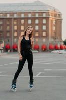 una foto completa de una mujer sonriente activa disfruta de paseos deportivos extremos en patines vestidos con ropa negra cómoda al aire libre aprende a patinar. concepto de recreación de ocio de personas