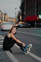 Outdoor shot of active slim woman poses on asphalt puts on rollerskates being in good mood spends free time riding rollerblades in urban place. Blurred city background. Hobby and recreation. photo