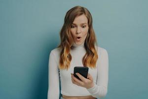 Shocked millennial girl looks at mobile phone with surprised face, eyes poped and mouth wide open photo