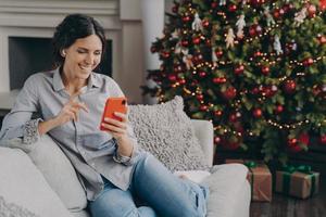 Happy female in earbuds holding smartphone, talking with family or friends online at Christmas time photo