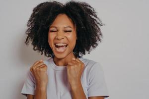 emocionada alegre chica de piel oscura aprieta los puños mientras cierra los ojos, exclama con victoria foto