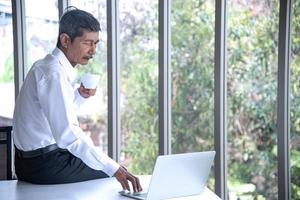 Work from home and social distancing concept. Asian business old man  working and drinking coffee at co-working space. Aging man using laptop meeting online. photo