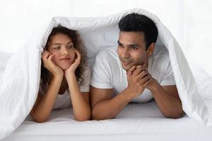 Funny couple lying, talking, hiding in white blanket on white bed  in the bedroom. Relation in family. Relaxing in the morning. photo